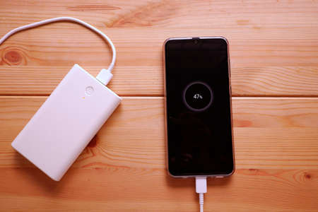 Smartphone is charging with backup battery or white power bank on the desk wooden tableの写真素材