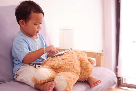 Asian boy about 2 years old watching and playing mobile phone sits on the sofa in the living room.の写真素材