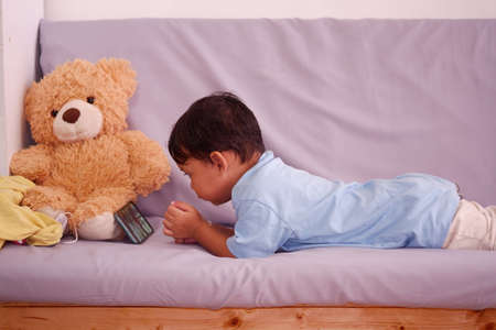 Asian boy about 2 years old, watching and playing mobile phone sits on the sofa in the living room. Ignoring the teddy bearの写真素材