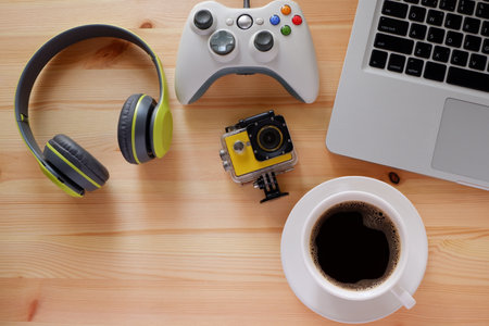 Wireless headphones and portable game consoles and other electronic gadgets on wooden desk with laptop computer with black coffee in white cupの写真素材