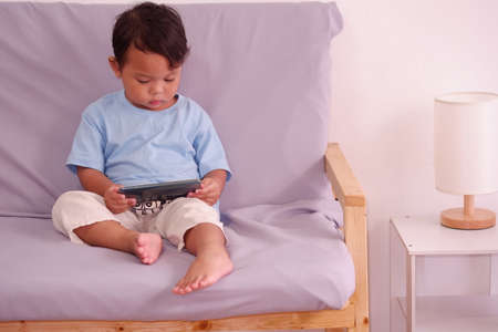 Cute asian child about 2 years old, watching and playing smartphone sits on the sofa in the living roomの写真素材
