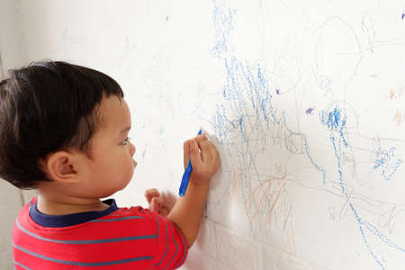 Cute asian boy in bright red striped t-shirt stands scribbling on white wall. with text copy space, Selective focusの写真素材