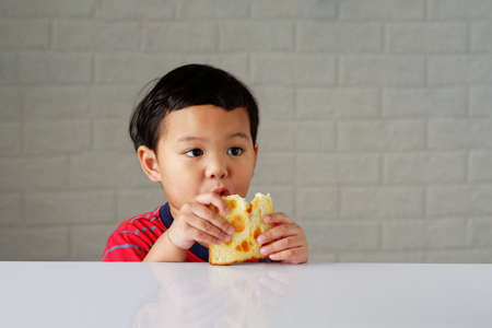 Cute little asian boy eating bread enjoying breakfast or snack on white tableの写真素材