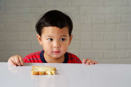 Cute little asian boy has an bored expression and is eating bread on a white dining tableの写真素材