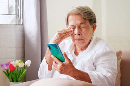 Elderly Asian woman with grey hair is using mobile phone and rubbing her eyes with her hand. Eye problems in the elderly, blurry vision, conceptの写真素材