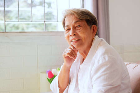 Elderly Asian woman with grey hair sits with her chin on the sofa with a happy smile on her face.の写真素材