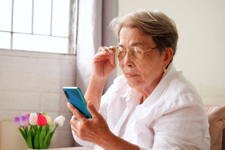 Elderly Asian woman with grey hair is using a mobile phone and holding her glasses. Eye problems in the elderly, blurry vision, conceptの写真素材