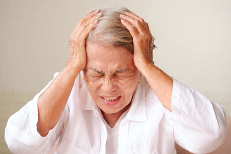 Elderly Asian woman with grey hair touches her painful head with both hands. Various illnesses of the elderly and problems that cause headaches concept.の写真素材