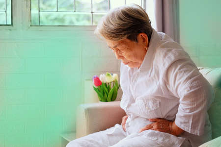 Elderly Asian woman with grey hair sits and holding her stomach in pain on the sofa, Aging society and Various illnesses of the elderly and health care concept, with copy space for text.の写真素材