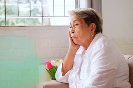 Elderly Asian woman with grey hair sits on her chin with a sad expression. Aging society Sad and lonely concept, with copy space for text.の写真素材