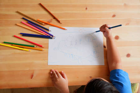 Cute Asian boy drawing happy family and home. with color pencils and white paper on wooden table, top viewの写真素材