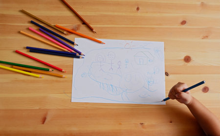 Cute Asian boy drawing happy family and home. with color pencils and white paper on wooden table, top viewの写真素材