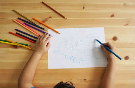 Cute Asian boy drawing happy family and home. with color pencils and white paper on wooden table, top viewの写真素材