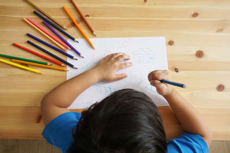 Cute Asian boy drawing happy family and home. with color pencils and white paper on wooden table, top viewの写真素材