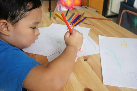 Cute Asian boy drawing with color pencils and white paper on wooden tableの写真素材