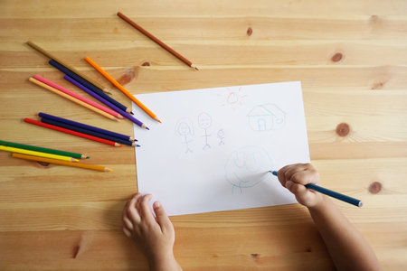 Cute Asian boy drawing happy family and home. with color pencils and white paper on wooden table, top viewの写真素材
