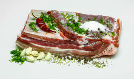 A piece of pork raw bacon with spices, greens, salt, garlic and chili pepper on a white backgroundの写真素材