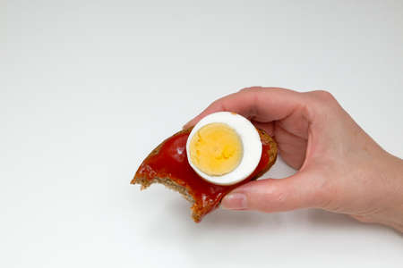 A woman's hand with a tinged rye bread sandwich, tomato sauce and two pieces of boiled egg on a white backgroundの写真素材