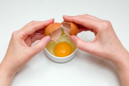 A woman's hand breaking a raw chicken egg into a white cup on a white backgroundの写真素材