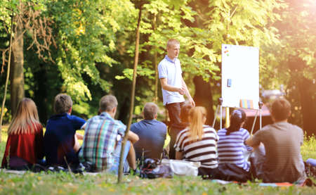 A group of young students in the park. View of a man gesticulating with his hands, standing against a defocused group of people sitting on the grass.の写真素材