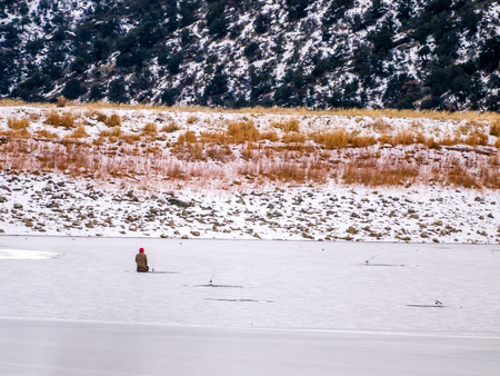 Solitary ice fisherman kneeling at a hole in the ice with fishing rodの写真素材