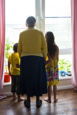 Grandmother and grandchildren are standing at a large window and look out on the street