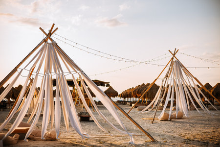 Boho tents made of wooden bars and white material on the beach. In the background, close to the sea, you can see straw umbrellas and sunbedsの写真素材