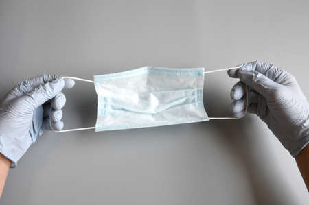 the doctor's hands protected by blue gloves hold a blue surgical mask on gray background, against...