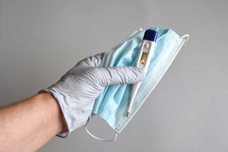 the doctor's hands protected by blue gloves hold a blue surgical mask and fever thermometer on gray...
