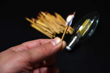 a man is holding a lit match with a light bulb and matches in the background. electricity supply problem concept. blackoutの写真素材