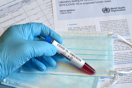 Florence, December 2021: New Omicron variant of the Covid 19 Virus. Doctor's hand in blue glove holds test tube with positive Omicron COVID-19 test blood sample with protective medical masks and documents on the background.のeditorial素材