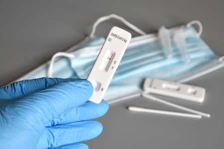 closeup of a healthcare worker, wearing blue surgical gloves, holding a covid-19 test device showing a negative result in his hand with protection mask on backgroundの写真素材