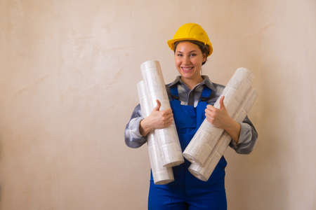 A girl in a blue construction workers jumpsuit and a yellow hard hat holds wallpaper in her hands,...