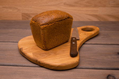 A loaf of fresh crusty bread sits on a wooden board next to a knife on the table. Pastries, flour...