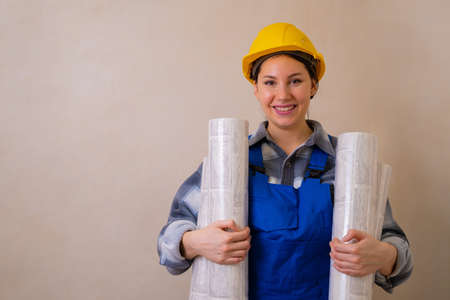 A beautiful young female construction worker in a blue uniform and hard hat holds two rolls of...