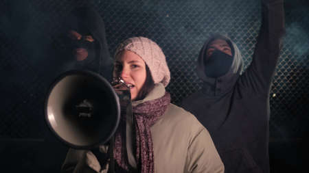 girl leader shouts slogans into a hand megaphone against the background of masked demonstrators....