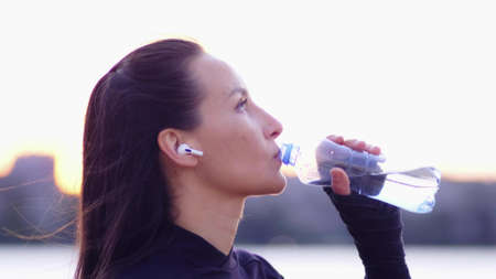 Asian woman with headphones in her ears drinks water from a plastic bottle