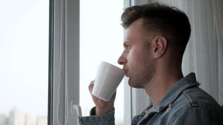 A young handsome man looks out the window and drinks coffee. Flavor tea in a cup. coffee break