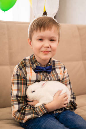 boy in a bunny ears headband holds a white rabbit in his arms and looks into the camera. Easter