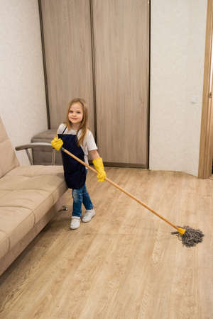 A young blonde little girl in yellow household rubber gloves and a blue apron smiles and cleans the floors in the roomの写真素材