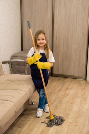 A young blonde little girl in yellow gloves and a blue apron smiles and holds a mop in her handsの写真素材