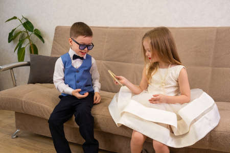A stylish little girl in a white dress and beads gives a bundle of banknotes to a boy in dark glasses. Family budgetの写真素材