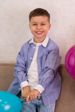 A little boy in a blue shirt and jeans is standing on the sofa, smiling and holding a balloonの写真素材