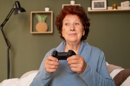 A modern elderly woman sits on a sofa in a room and plays a computer game on a gamepad. Hobbies and...