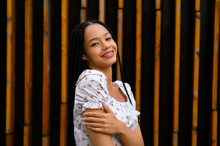 Portrait of a young pretty afro woman with pigtails who smiles, looks at the camera and enjoys herself, posing half sideways and hugging her handの写真素材