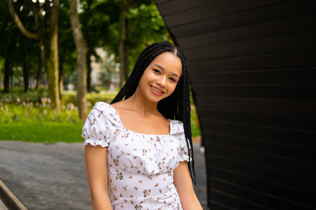 Portrait of an African young woman standing on the street in a park in a summer dress, smiling and looking at the cameraの写真素材