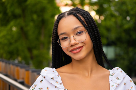 Close-up portrait of a young attractive African woman wearing glasses standing on a bridge on the street in a park in summer. Studentの写真素材