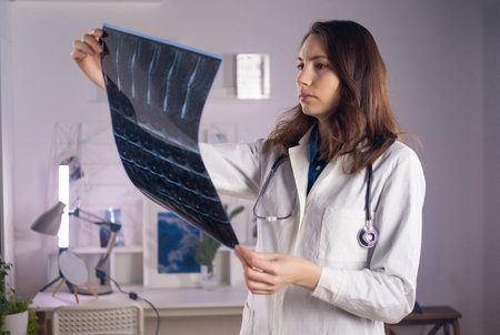 A young adult white female doctor in a medical mask and a white coat holds a CT scan in her hands and looks at it carefullyの写真素材