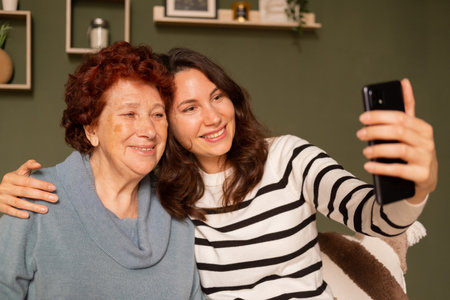 Portrait of a modern happy grandmother and granddaughter sitting on the sofa during the day in the living room. A young woman hugs an elderly woman by the shoulder and they take a selfie with a smileの写真素材