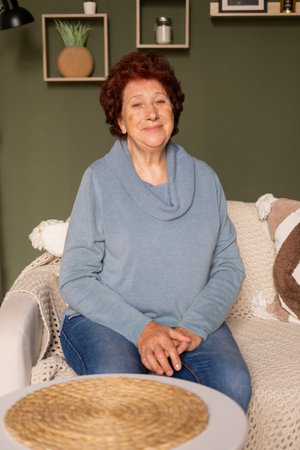 Portrait of an elderly woman with blue eyes and red hair looks at the camera in a room sitting on a sofaの写真素材